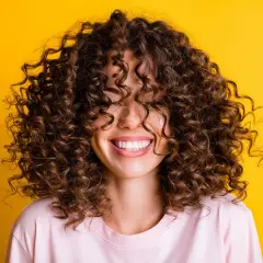Mujer sonriendo con cabello rizado castaño sobre fondo amarillo.