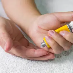 Manos aplicando crema de un tubo amarillo en la palma de otra mano.