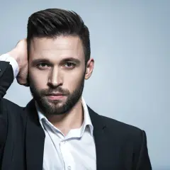 Hombre con un peinado con tupe, barba y traje negro, tocándose la cabeza.