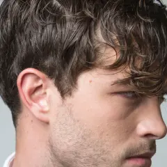 El pelo hombre, oscuro y ondulado, cae sobre su rostro. Se aprecian su oreja y parte de su barba incipiente.