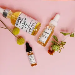 Tres frascos de aceite facial "Clarify Me" con escualeno, lavanda y neroli, rodeados de flores secas sobre fondo rosa.