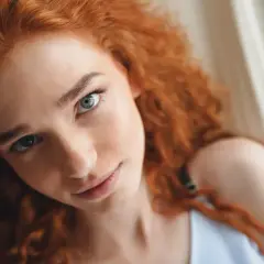 Primer plano de una joven con cabello pelirrojo natural y ojos claros, recostada en una hamaca.