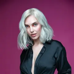 Mujer con cabello rubio ceniza platino, ojos azules y camisa negra abierta, sobre fondo rosa.