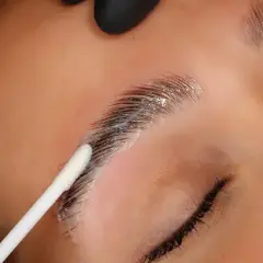 Aplicación de producto para que es el laminado de cejas. Un hisopo blanco aplica una crema sobre las cejas oscuras y peinadas.