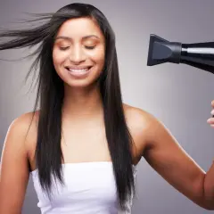 Mujer sonríe mientras se seca el pelo fino y escaso con un secador negro.