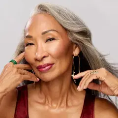 Mujer con cabello gris, con un elegante efecto de grey blending, sonríe con labios rojos y joyas doradas y verdes.