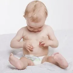 Bebé con piel atópica, sentado con pañal, examinando su barriguita con curiosidad.