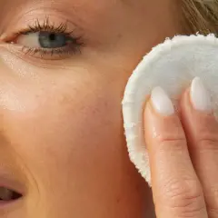 Rostro de mujer rubia con piel luminosa, aplicándose un disco desmaquillante. Su piel luce sin imperfecciones, sin rastro de milium.