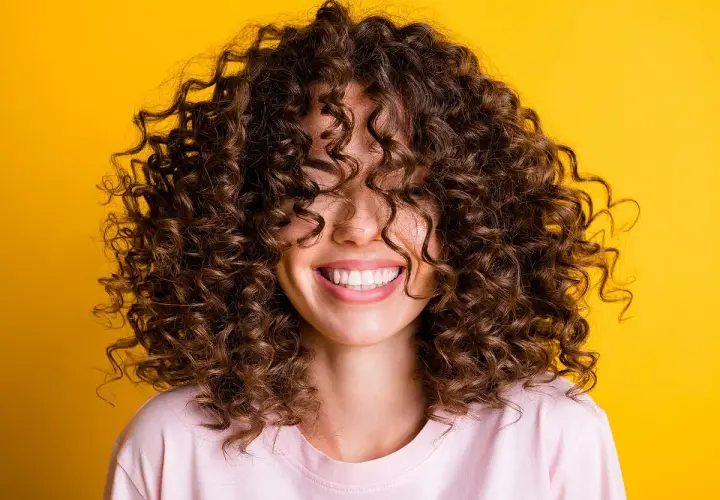 Mujer sonriendo con cabello rizado castaño sobre fondo amarillo.
