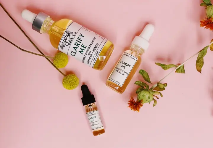 Tres frascos de aceite facial "Clarify Me" con escualeno, lavanda y neroli, rodeados de flores secas sobre fondo rosa.