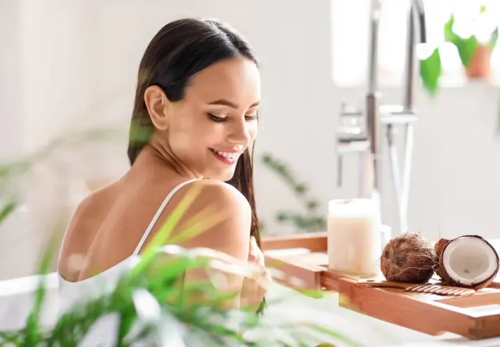 Mujer sonriente en la bañera con cocos y un frasco de aceite de coco, mostrando para qué sirve el aceite de coco para el cabello.
