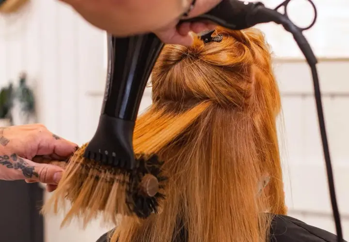 Manos con tatuajes secan y peinan cabello pelirrojo con secador y cepillo redondo. El brushing que es, es una técnica de estilismo.