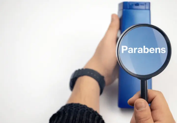 Lupa amplía la palabra "Parabens" sobre un frasco azul, indicando la búsqueda de ingredientes en productos.