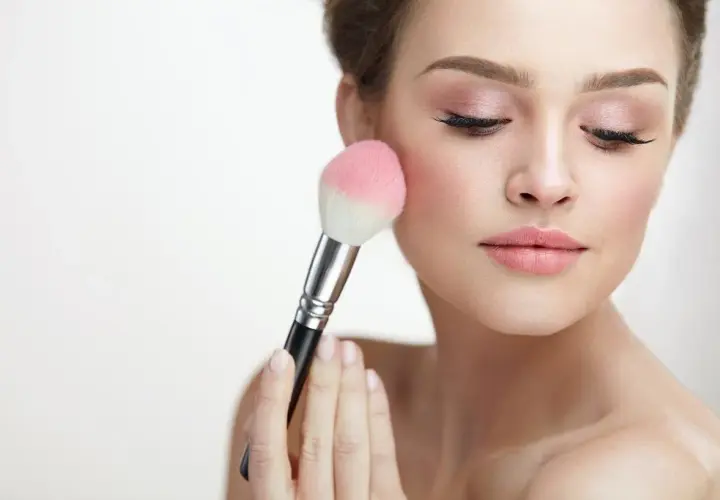 Mujer aplicando rubor en sus mejillas con una brocha. El tono rosado del rubor realza su piel, creando un look natural y radiante.