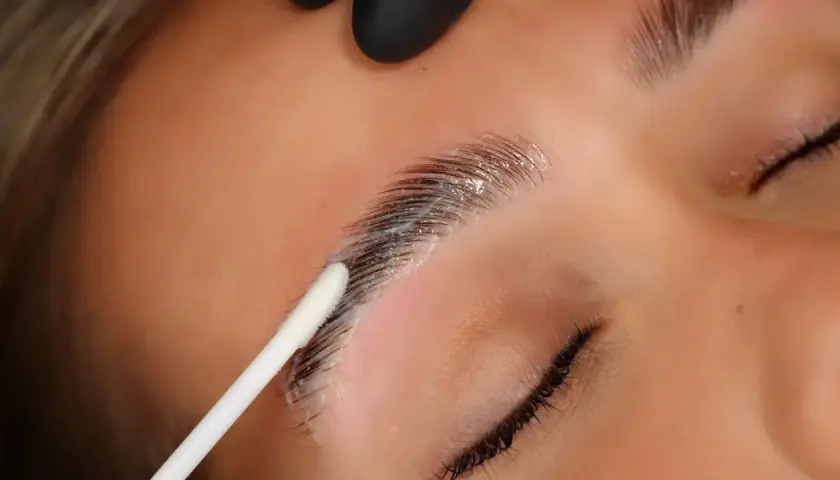 Aplicación de producto para que es el laminado de cejas. Un hisopo blanco aplica una crema sobre las cejas oscuras y peinadas.