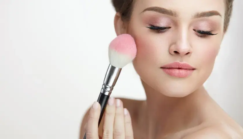 Mujer aplicando rubor en sus mejillas con una brocha. El tono rosado del rubor realza su piel, creando un look natural y radiante.