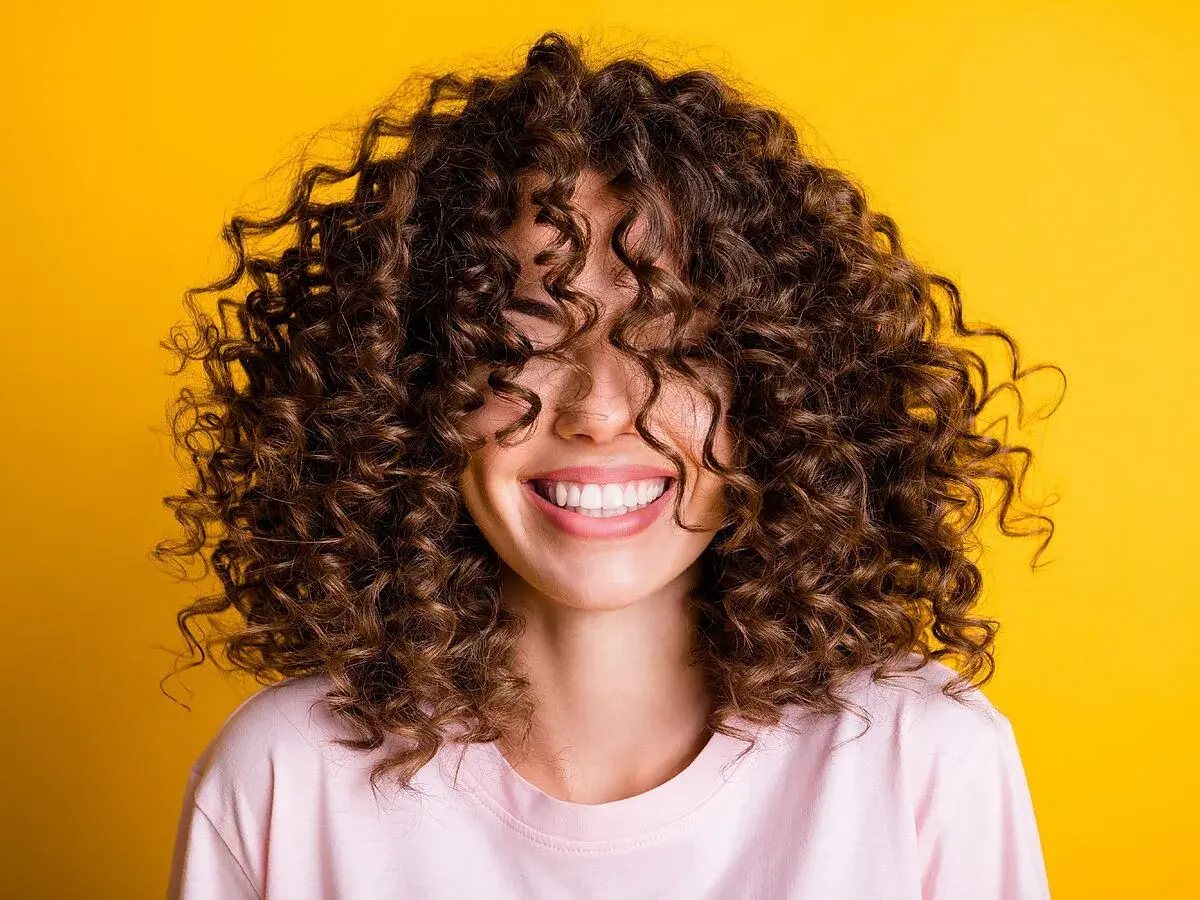 Mujer sonriendo con cabello rizado castaño sobre fondo amarillo.