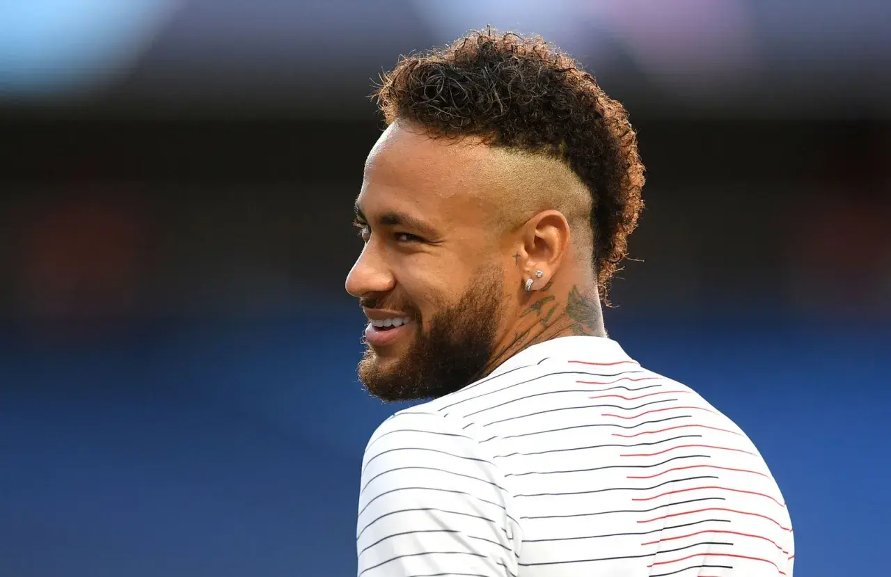 Sonrisa de Neymar con su característico peinado mohicano, luciendo una camiseta a rayas blancas y rojas.