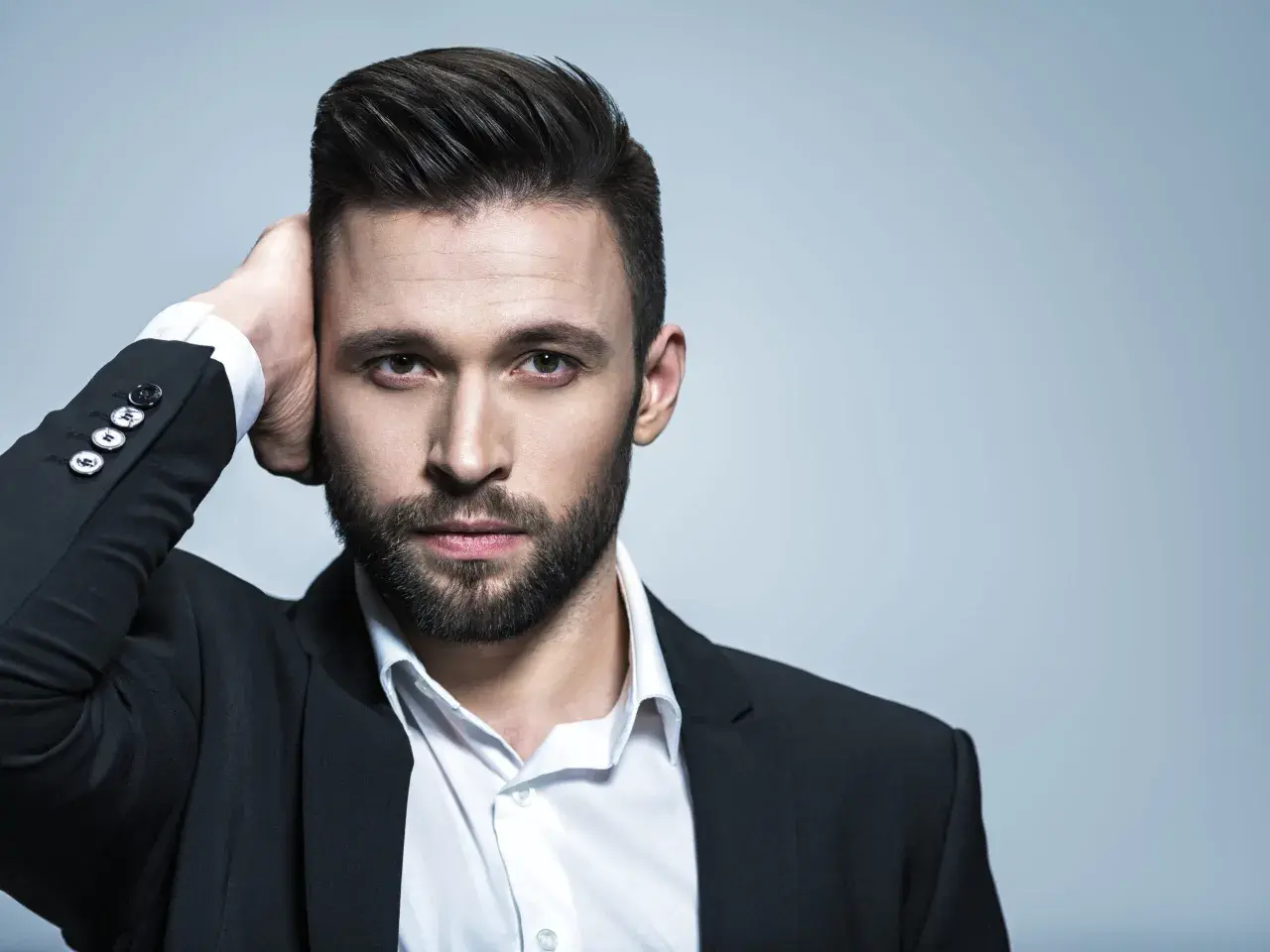 Hombre con un peinado con tupe, barba y traje negro, tocándose la cabeza.