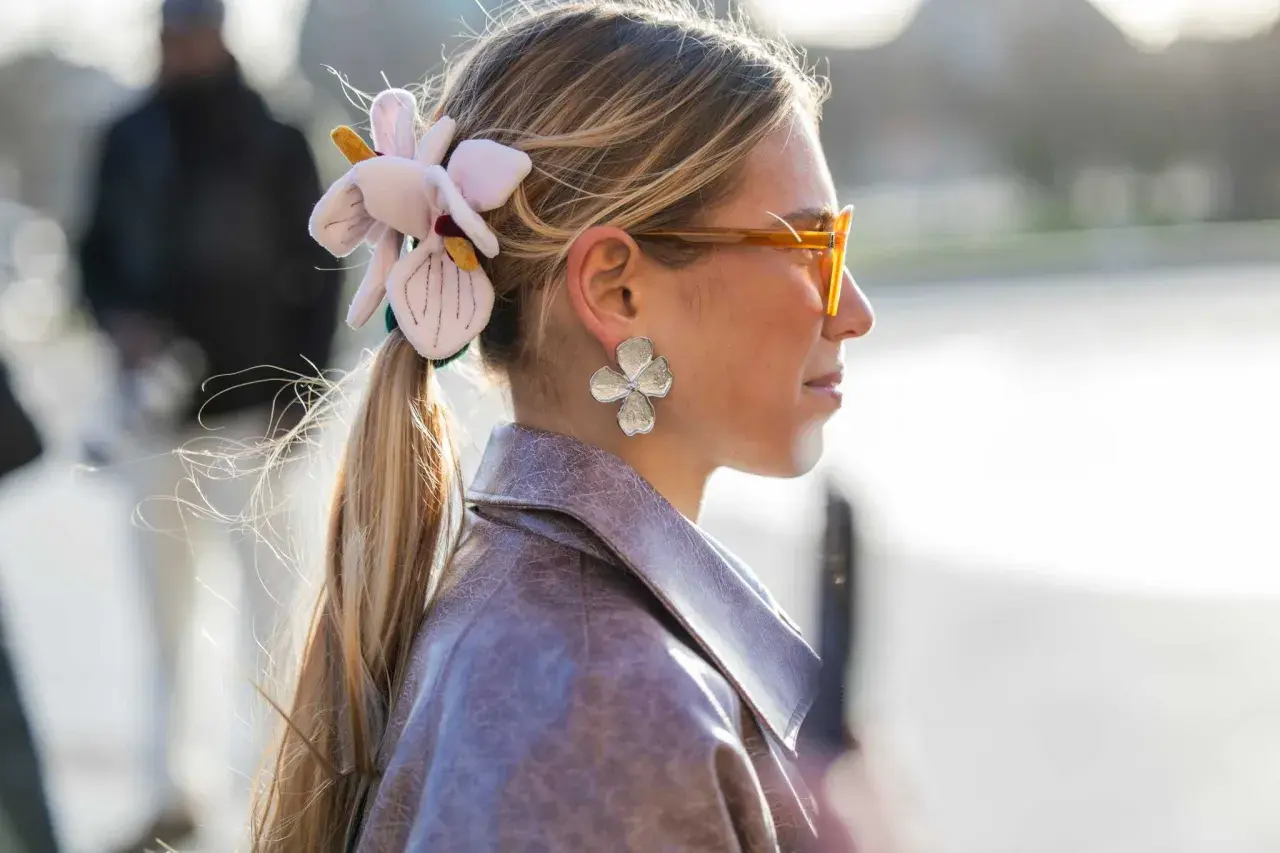 Mujer con coleta adornada con una flor de tela y pendientes de pétalos, luciendo gafas naranjas y una chaqueta de cuero.