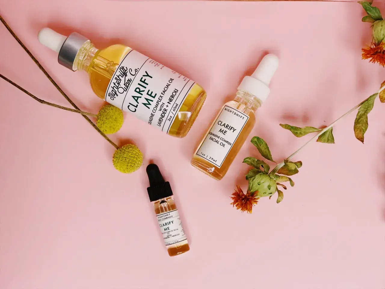 Tres frascos de aceite facial "Clarify Me" con escualeno, lavanda y neroli, rodeados de flores secas sobre fondo rosa.