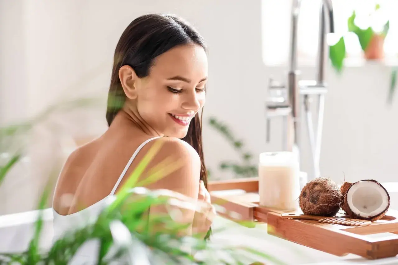 Mujer sonriente en la bañera con cocos y un frasco de aceite de coco, mostrando para qué sirve el aceite de coco para el cabello.