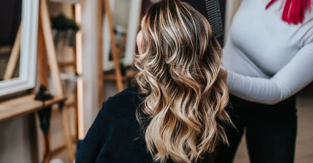 Mujer con cabello castaño y mechas rubias ondulado, visto desde atrás en un salón de belleza.
