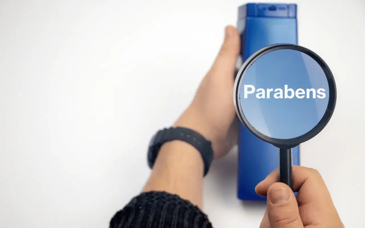 Lupa amplía la palabra "Parabens" sobre un frasco azul, indicando la búsqueda de ingredientes en productos.