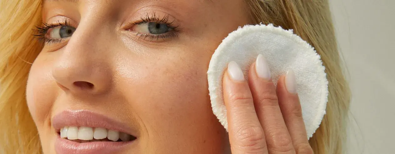 Rostro de mujer rubia con piel luminosa, aplicándose un disco desmaquillante. Su piel luce sin imperfecciones, sin rastro de milium.