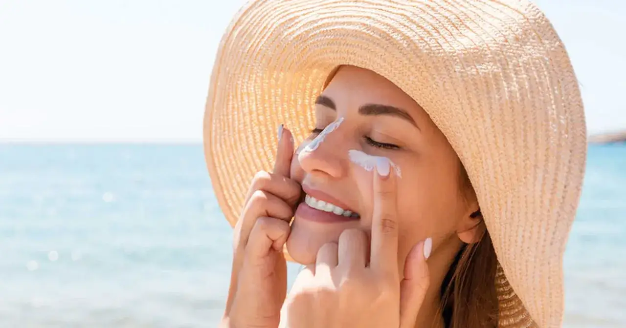 Mujer con sombrero de paja aplicándose protector solar en la cara, previniendo el melasma en un día soleado en la playa.