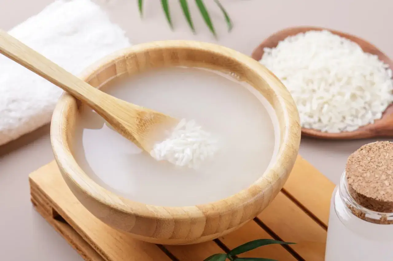 Un tazón de madera contiene agua de arroz con una cuchara de bambú. Arroz blanco en una cuchara de madera y una toalla blanca al lado.