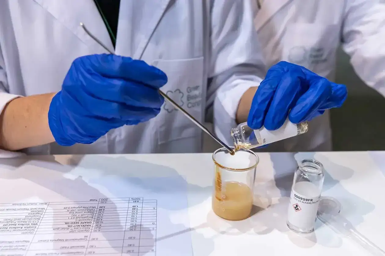 Manos con guantes azules añaden líquido a un vaso de precipitados. En la mesa, una lista de ingredientes incluye phenoxyethanol.