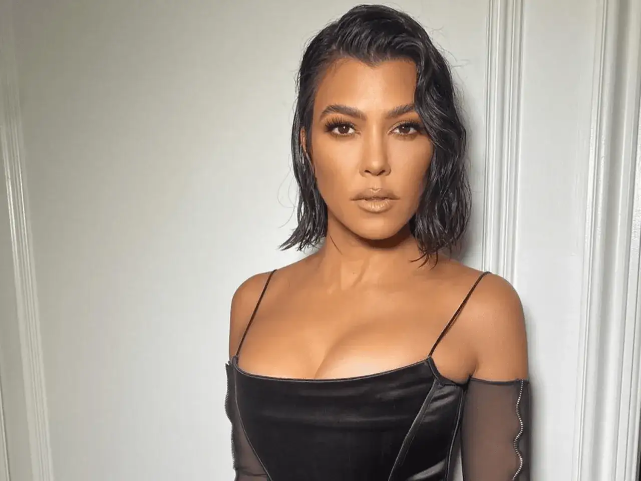 Mujer con cabello oscuro y ondulado, labios dorados y un vestido negro con escote pronunciado. Su peinado resalta un sutil pico de viuda.