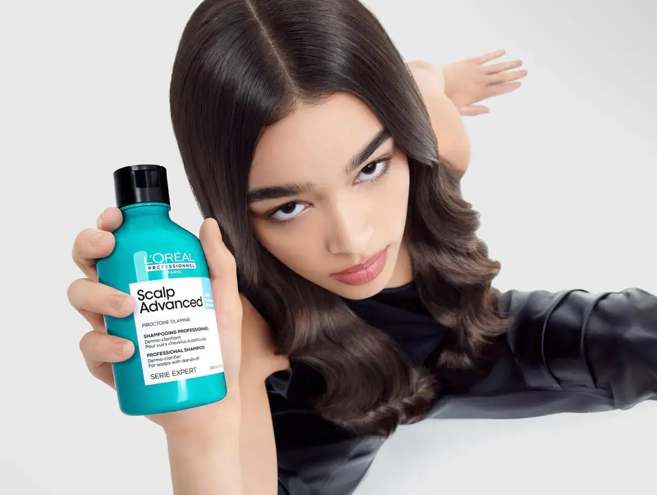 Mujer con cabello oscuro sostiene un frasco de champú L'Oréal Scalp Advanced, ideal para el cuero cabelludo irritado.