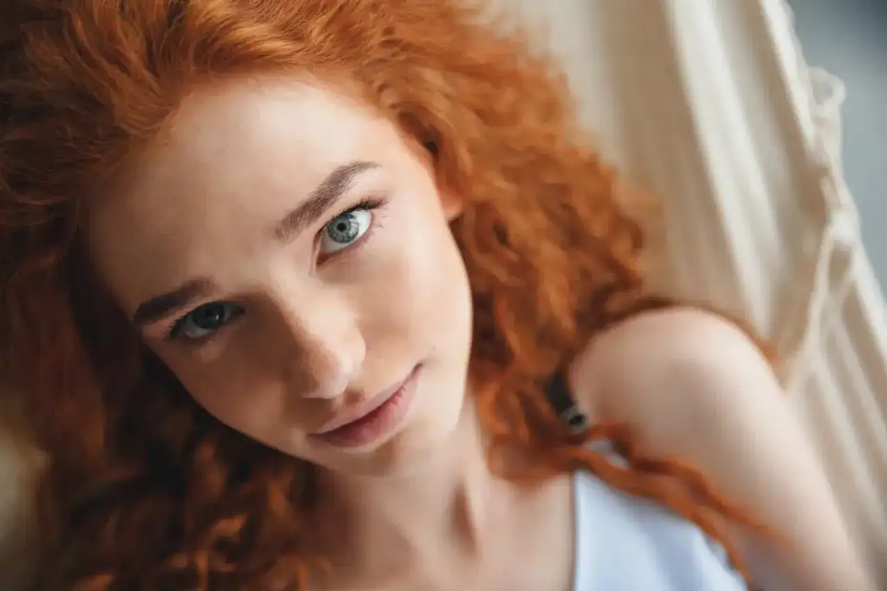 Una joven pelirroja con ojos azules penetrantes mira a la cámara, su cabello rizado enmarca su rostro.