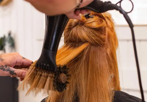 Manos con tatuajes secan y peinan cabello pelirrojo con secador y cepillo redondo. El brushing que es, es una técnica de estilismo.